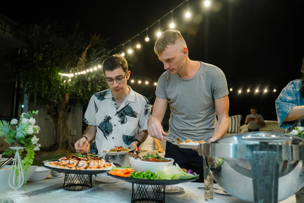 Men serving food at outdoor buffet with string lights.